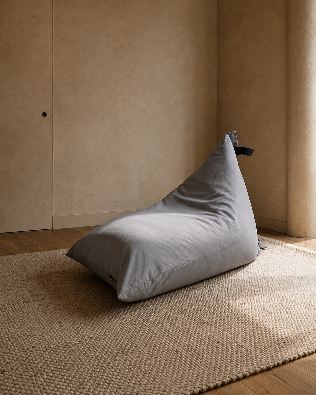 A light gray fabric bean bag chair resting on a textured beige woven rug in a minimalist room with warm beige walls and wooden flooring.