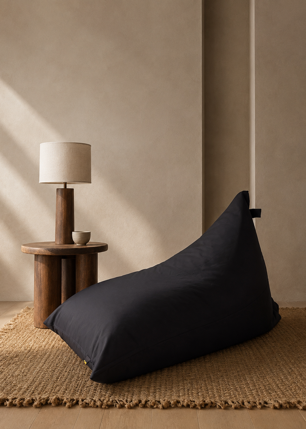 A black bean bag chair placed on a woven jute rug next to a wooden side table with a table lamp in a minimalist, warm-toned room.