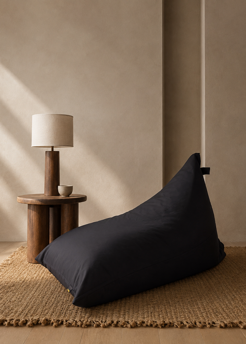 A black bean bag chair placed on a woven jute rug next to a wooden side table with a table lamp in a minimalist, warm-toned room.