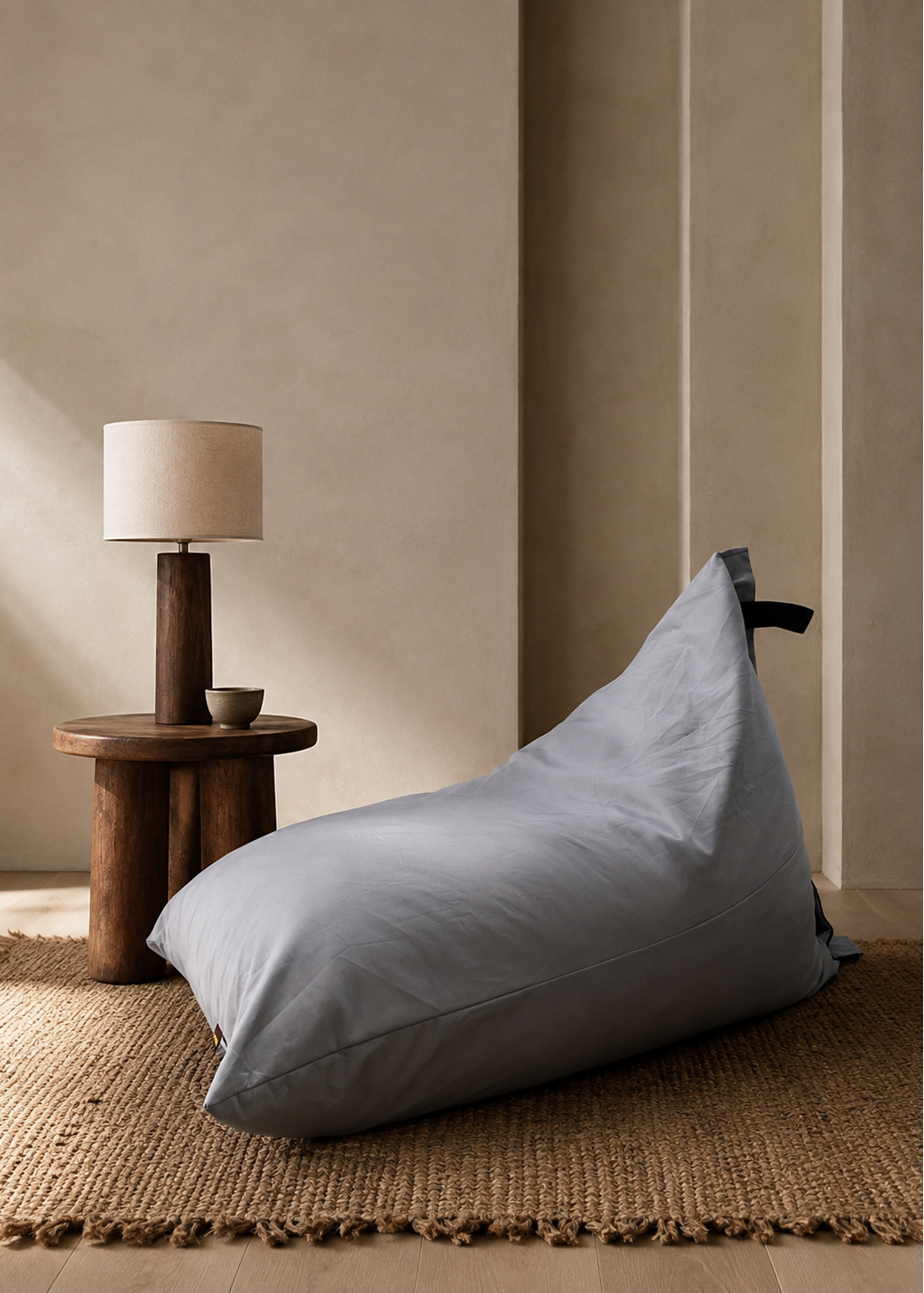 A large light gray bean bag chair sits on a woven jute rug in a minimalist room, next to a small wooden side table with a table lamp.