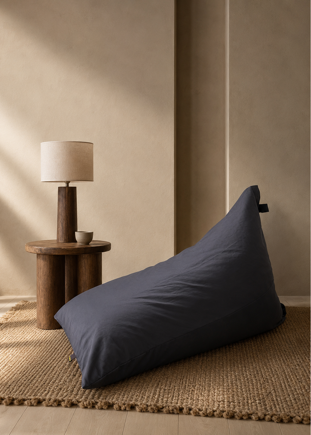 A large dark slate blue bean bag chair sits on a woven jute rug in a minimalist, warm-toned room next to a wooden side table with a lamp.