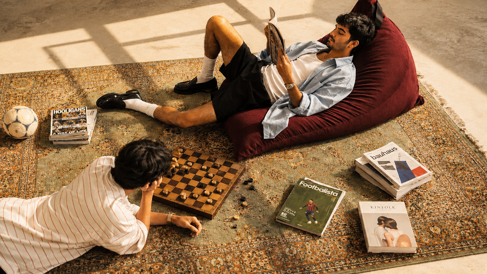 Two men relax in a stylishly decorated room. One man lies on a large burgundy bean bag chair reading a magazine, while the other lies on a patterned rug playing chess.