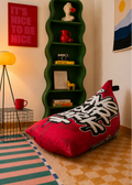 A red bean bag chair with bold black and white graffiti-style lettering sits on a checkered floor in a styled room. A green wavy bookshelf, a red floor lamp, and colorful wall art are visible in the background.