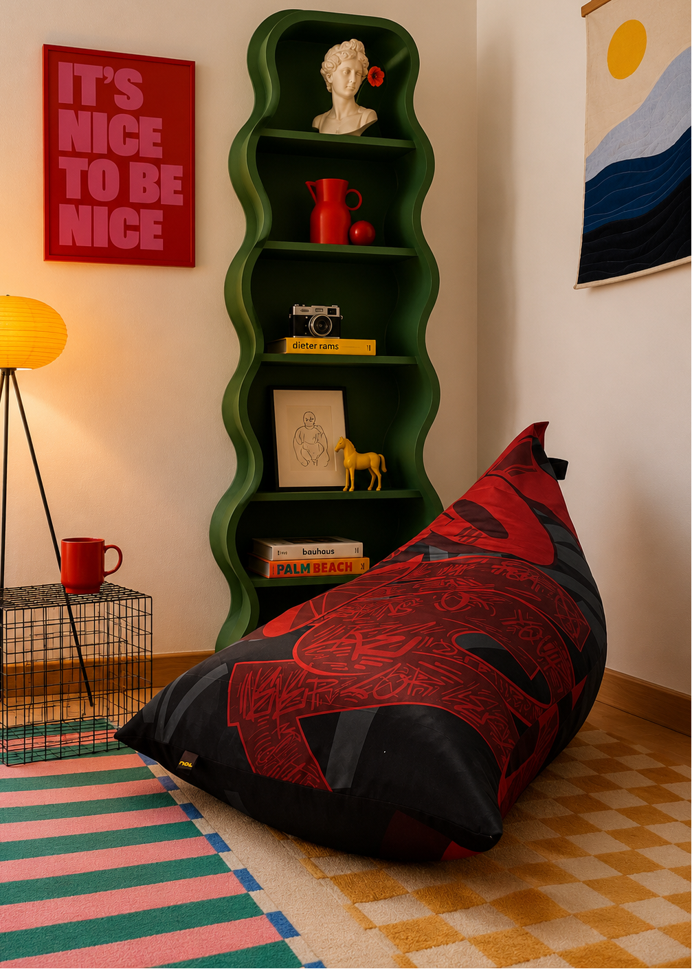 A large bean bag chair with a red and black ornamental pattern sits on a checkered floor in a styled room. A tall green wavy shelf stands behind it holding decorative objects, next to a red poster and a yellow floor lamp.
