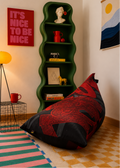 A large bean bag chair with a red and black ornamental pattern sits on a checkered floor in a styled room. A tall green wavy shelf stands behind it holding decorative objects, next to a red poster and a yellow floor lamp.