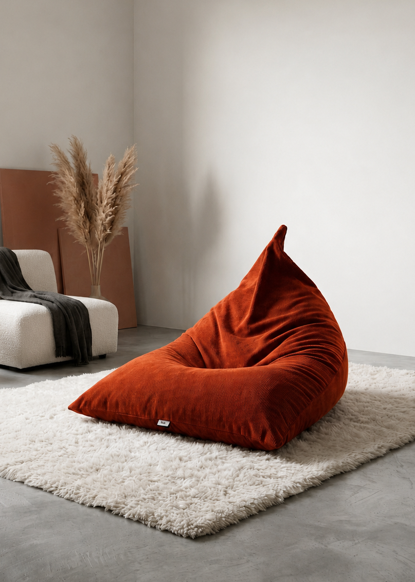 A rust-orange velvet bean bag chair placed on a white shag rug in a minimalist modern room with a white armchair, wooden side table, and dried pampas grass.