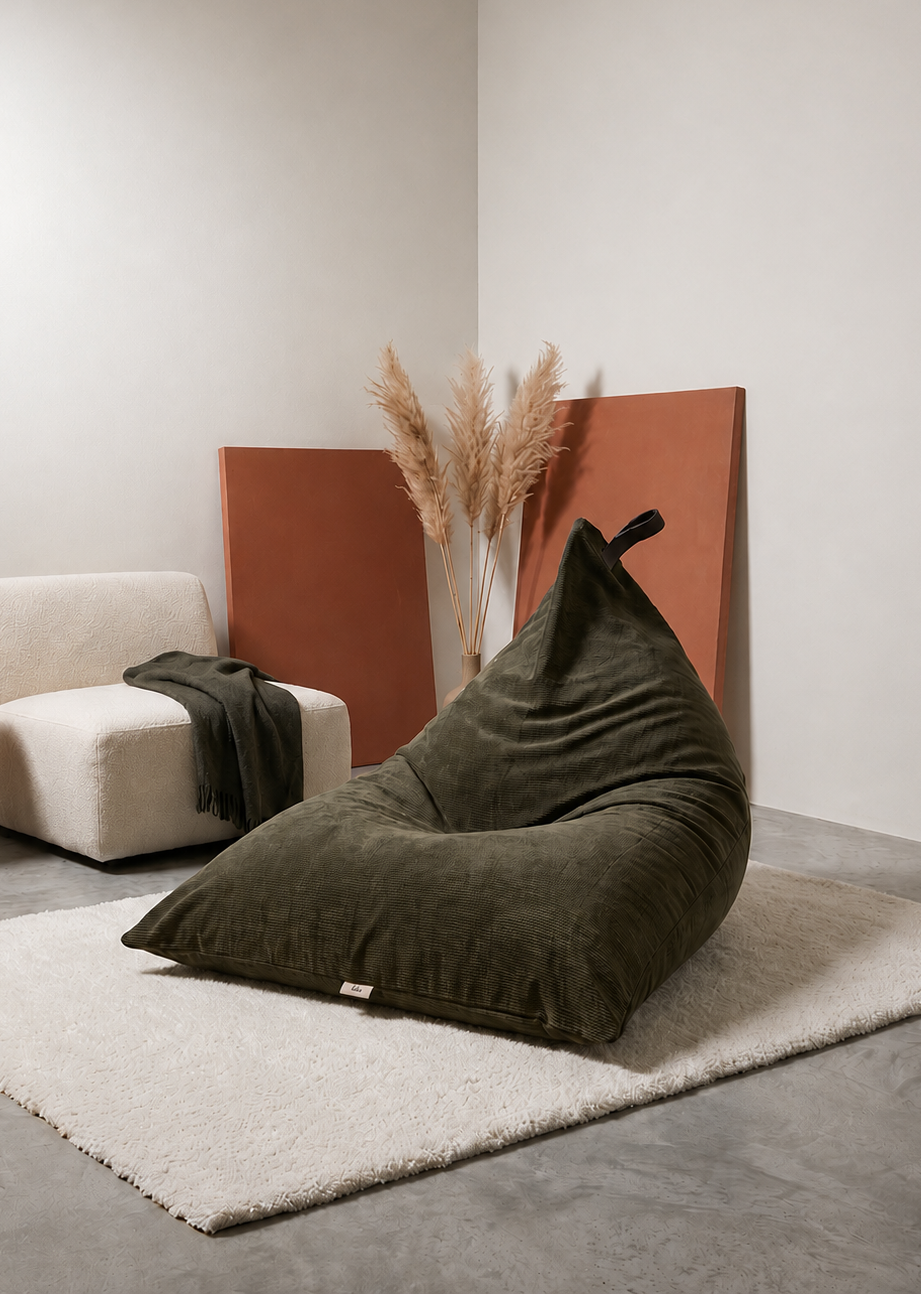 An olive green velvet bean bag chair styled in a minimalist room corner with a white armchair, a small wooden side table, a white rug, and pampas grass decor.