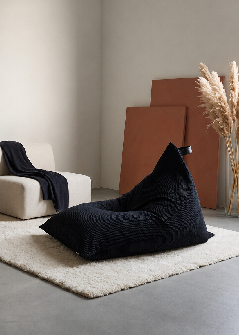 A large navy bean bag chair sits on a cream shag rug in a minimalist, modern room with a small cream armchair, a wooden side table, and pampas grass in the background.
