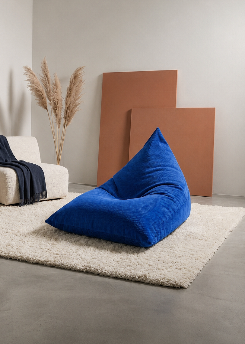 A bright blue corduroy bean bag chair placed on a white shag rug in a minimalist modern living room setting with a white armchair, navy throw blanket, wooden side table, and terracotta panels on the wall.