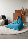 A teal blue bean bag chair placed on a round beige shag rug in a minimalist living room setting with a white armchair, black throw blanket, brown side table, and dried pampas grass.