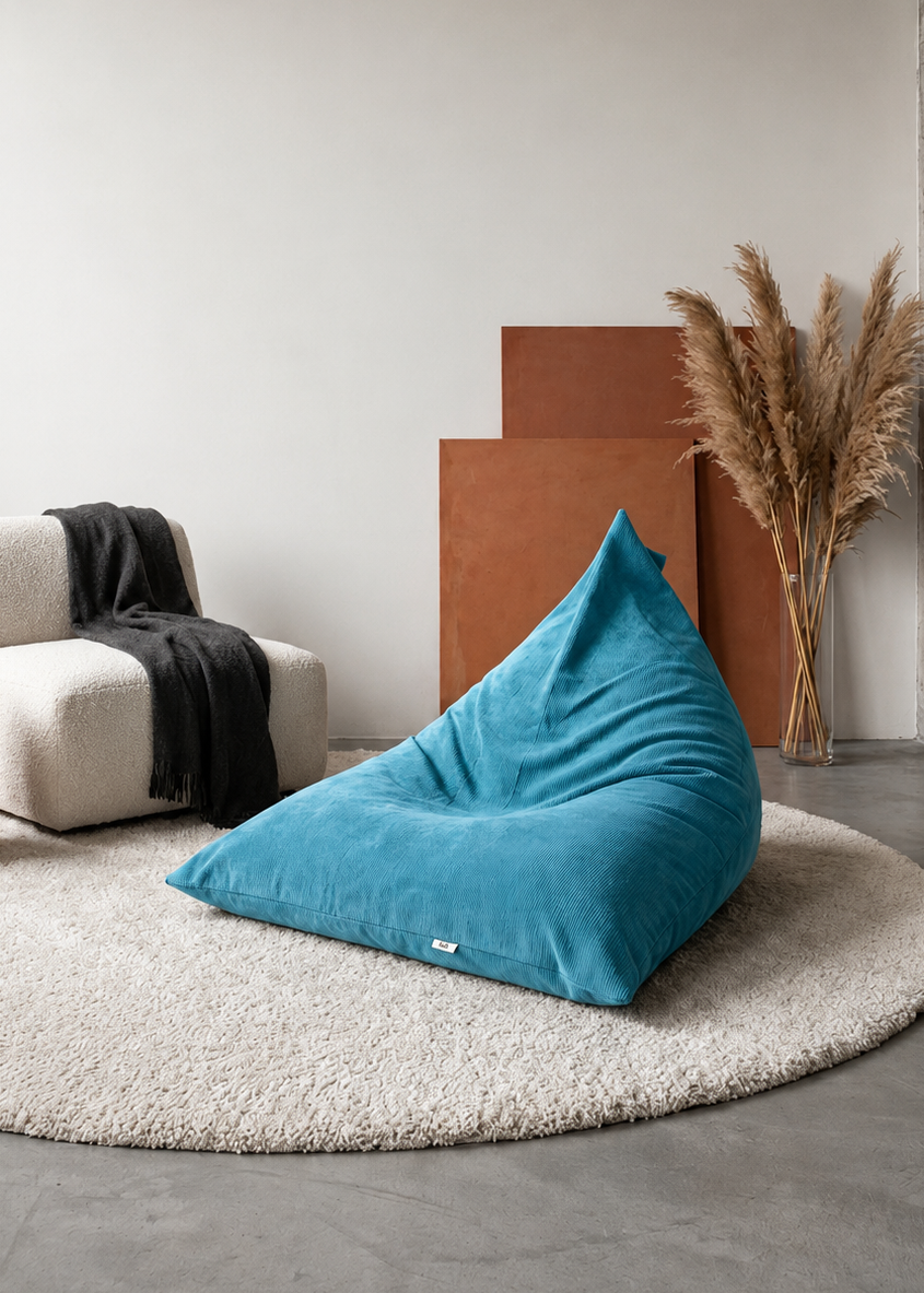 A teal blue bean bag chair placed on a round beige shag rug in a minimalist living room setting with a white armchair, black throw blanket, brown side table, and dried pampas grass.
