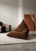A large brown corduroy bean bag chair sits on a white shag rug in a minimalist modern living room. In the background, a white armchair with a brown throw blanket and a small wooden side table are visible next to a terracotta-colored panel and pampas grass.