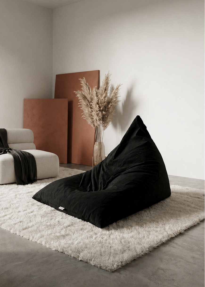 A large black fabric bean bag chair sits on a white shag rug in a minimalist, modern living room setting with a white armchair, draped black throw, and terracotta panels in the background.