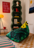 A green and yellow bean bag chair with a tropical leaf pattern sits on a checkered floor in a styled room with a green wavy bookshelf, a red framed poster, and a yellow floor lamp.