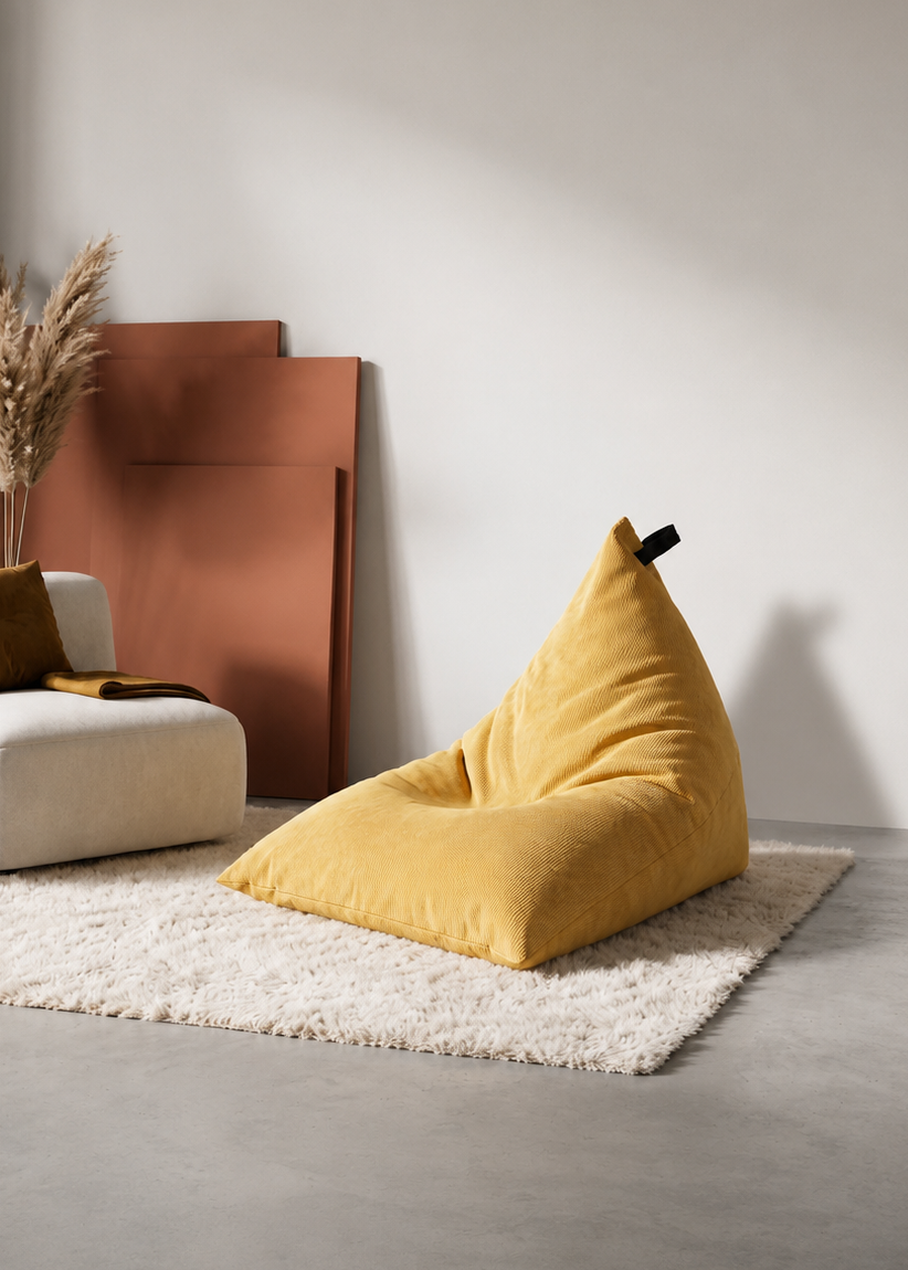 A mustard yellow bean bag chair sits on a white shag rug in a minimalist living room setting next to a white armchair and a small wooden stool.