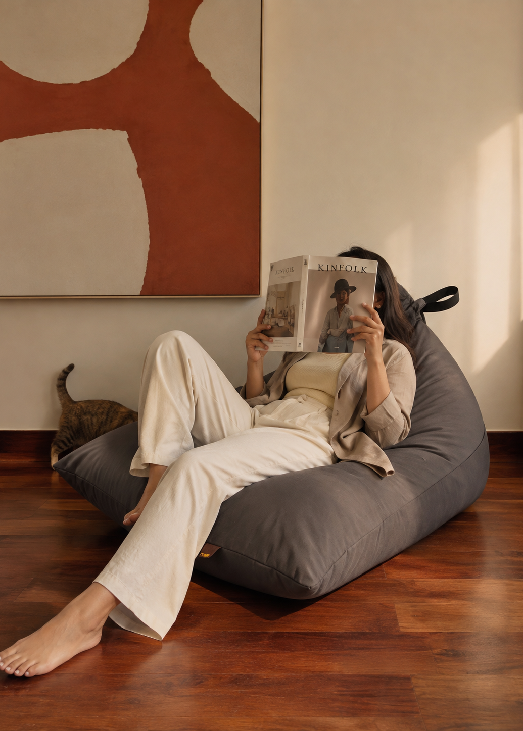 A person lounges on a dark gray bean bag chair in a cozy room with hardwood floors, reading a magazine. A cat is visible on the floor nearby, and a large abstract wall art piece hangs on the wall behind.