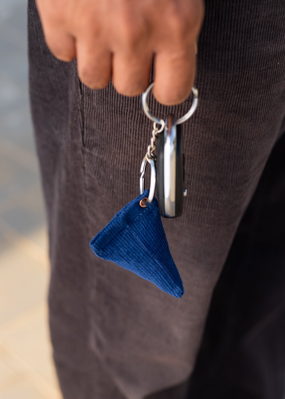 A close-up of a person's hand holding a silver metal keyring with a small blue corduroy triangular keychain bean bag, worn over dark brown corduroy pants.