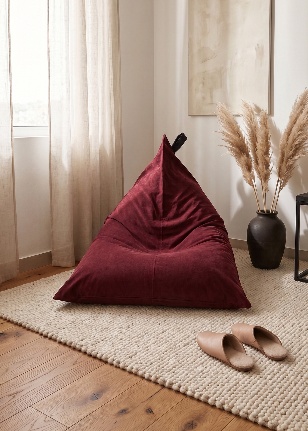 A triangular deep burgundy velvet bean bag chair placed on a woven jute rug in a modern, sunlit room with wooden floors, beige curtains, a black side table, and tan slippers.