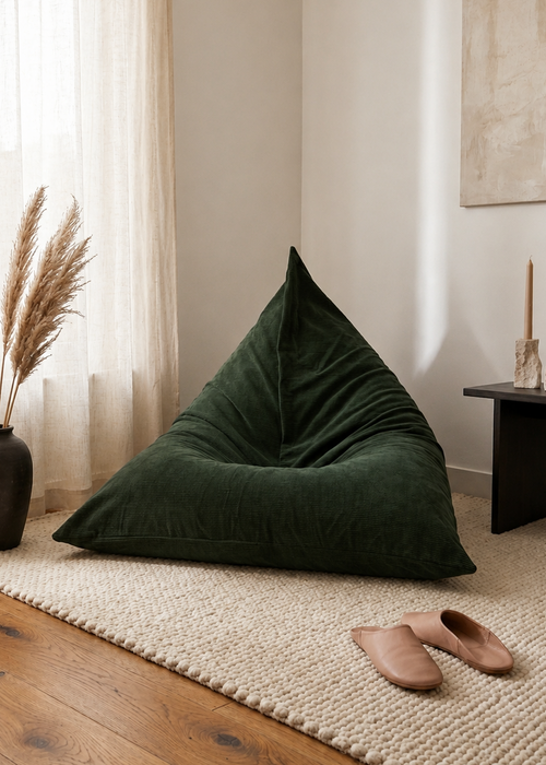 A dark green corduroy bean bag chair placed on a woven beige rug in a styled room corner with wood flooring, sheer curtains, a vase of pampas grass, and a side table.