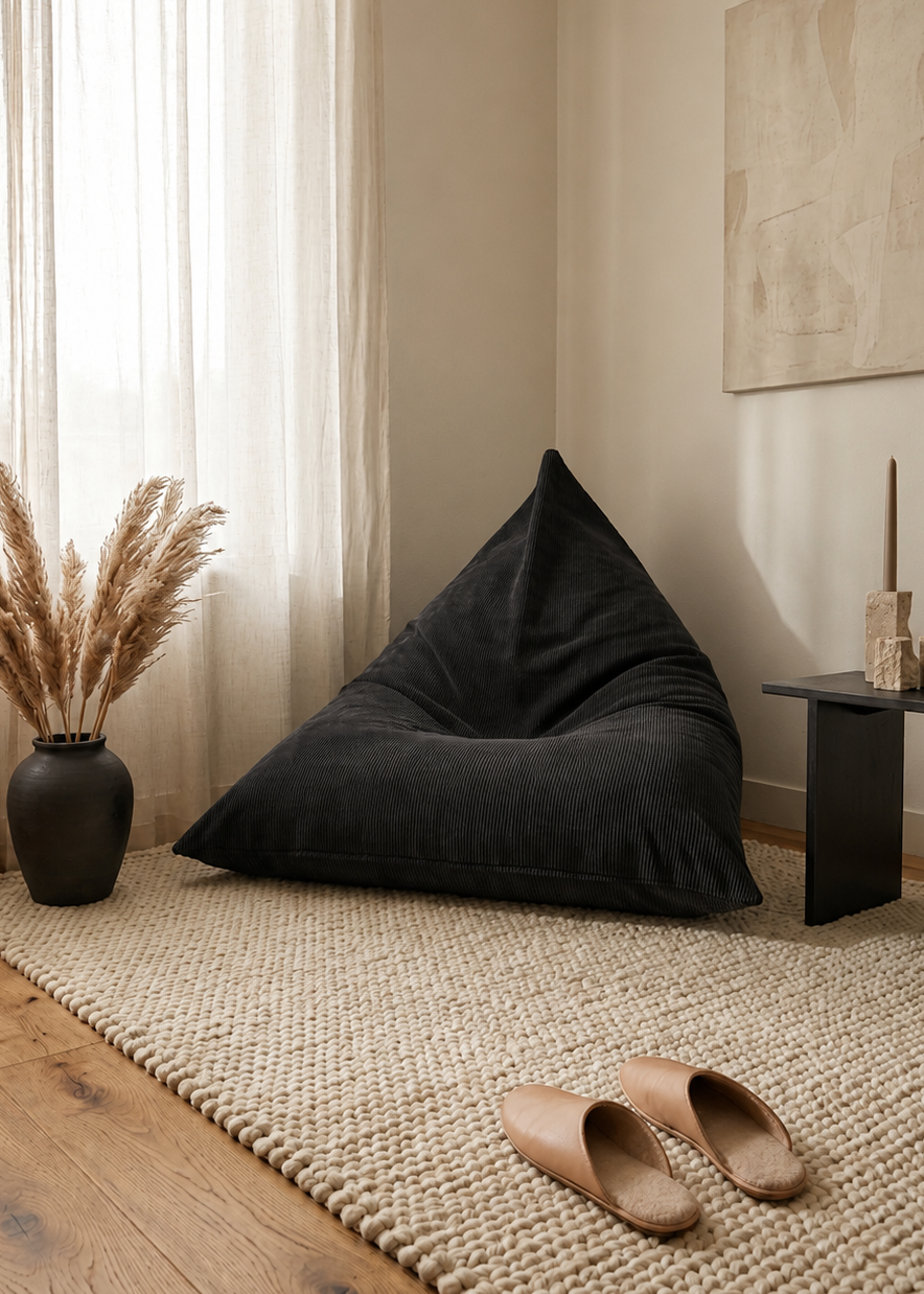 A large black bean bag chair in a cozy, neutral-toned room with a woven rug, wooden floor, sheer curtains, and a vase of pampas grass.
