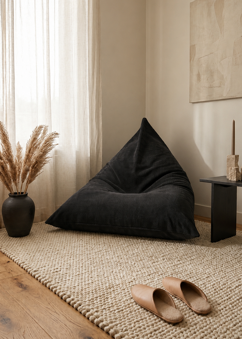 A large black bean bag chair in a cozy, neutral-toned room with a woven rug, wooden floor, sheer curtains, and a vase of pampas grass.