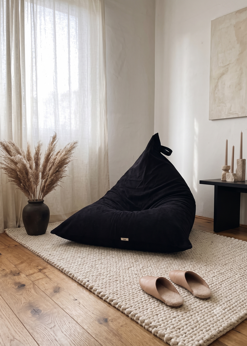 A navy bean bag chair sits on a chunky cream woven rug in a bright, modern room with light wood floors, sheer curtains, a vase of pampas grass, and a pair of tan slip-on slippers.