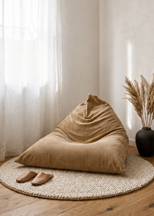 A large beige corduroy bean bag chair sits on a round woven jute rug in a bright, minimalist room with light wood floors, sheer white curtains, and a black side table with dried pampas grass.