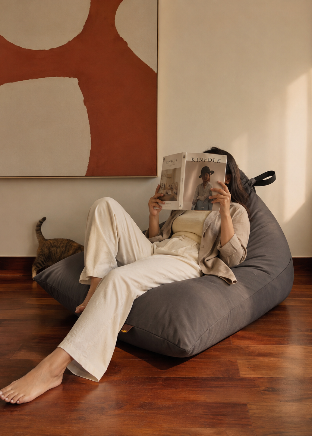A person lounges on a dark gray bean bag chair in a cozy room with hardwood floors, reading a magazine. A cat is visible on the floor nearby, and a large abstract wall art piece hangs on the wall behind.