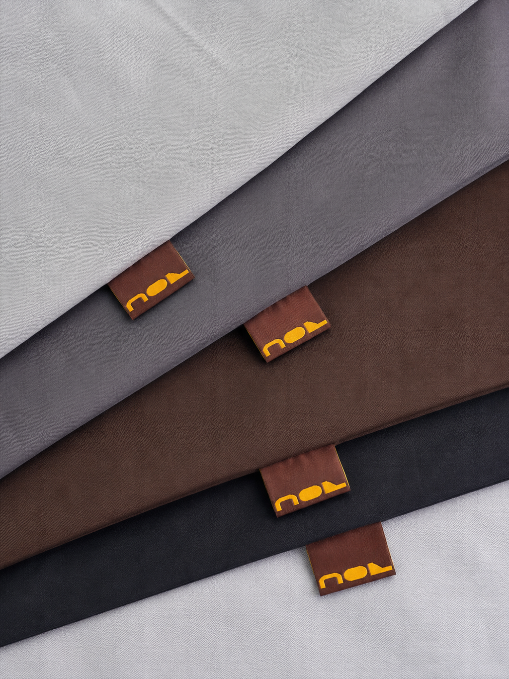 Four folded fabric sheets or blankets in light gray, charcoal gray, medium brown, and black, arranged diagonally. Each fabric has a small brown leather-like tag with a yellow-orange logo that appears to read 'noa'.