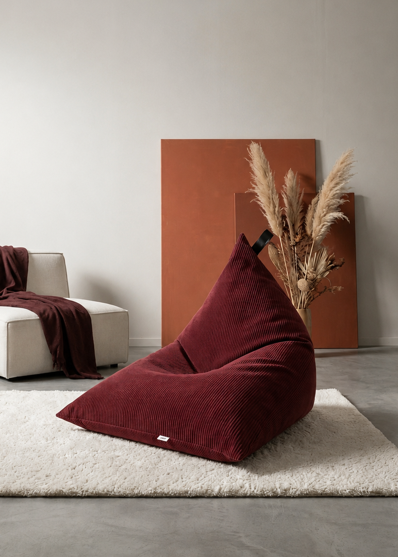 A deep burgundy corduroy bean bag chair sits on a white shag rug in a minimalist, modern living room setting with a white armchair, wooden stool, and pampas grass decor.
