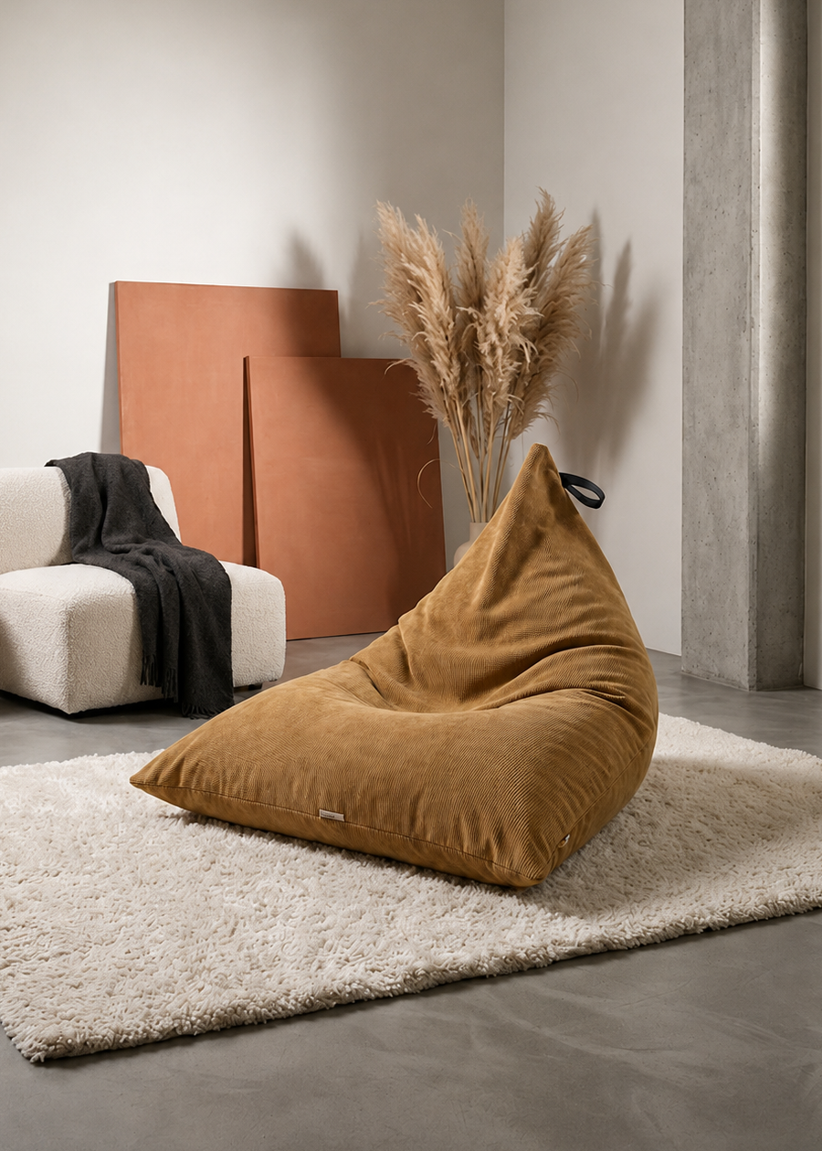 A mustard brown corduroy bean bag chair placed on a cream shag rug in a minimalist, modern room setting with a white armchair, wooden panels, and dried pampas grass.